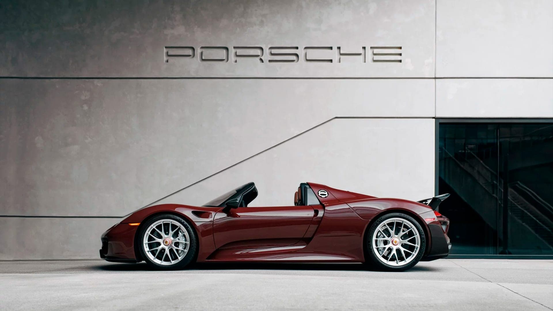 Burgundy Porsche 918 Spyder in side profile against a concrete wall